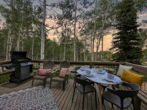 A cozy outdoor deck with a grill, dining table set for four, and chairs surrounded by trees at sunset.